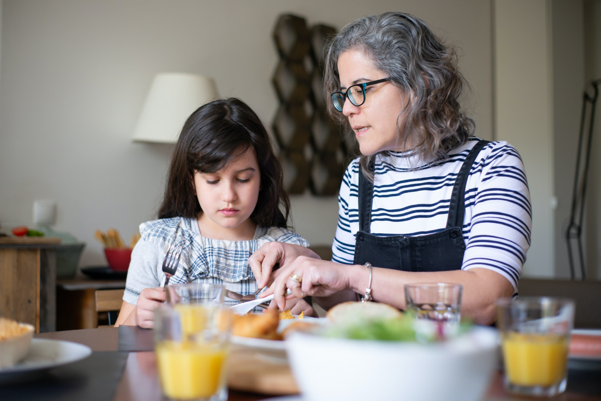 Woman helps a young girl cut food, in front of them is a table with glasses and a bowl of food