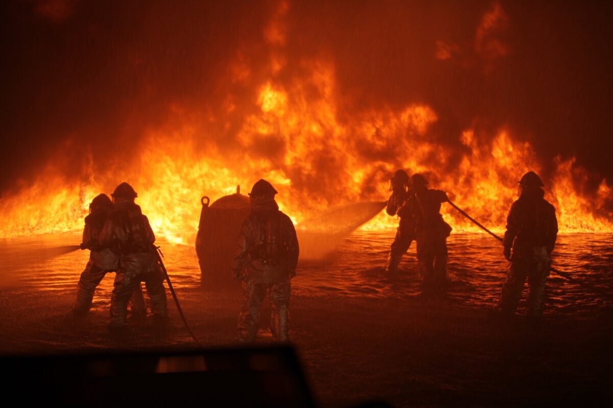 Backlit firefighters battle a large fire with hoses.